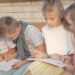 4 children sitting on bench outside school writing in notebooks